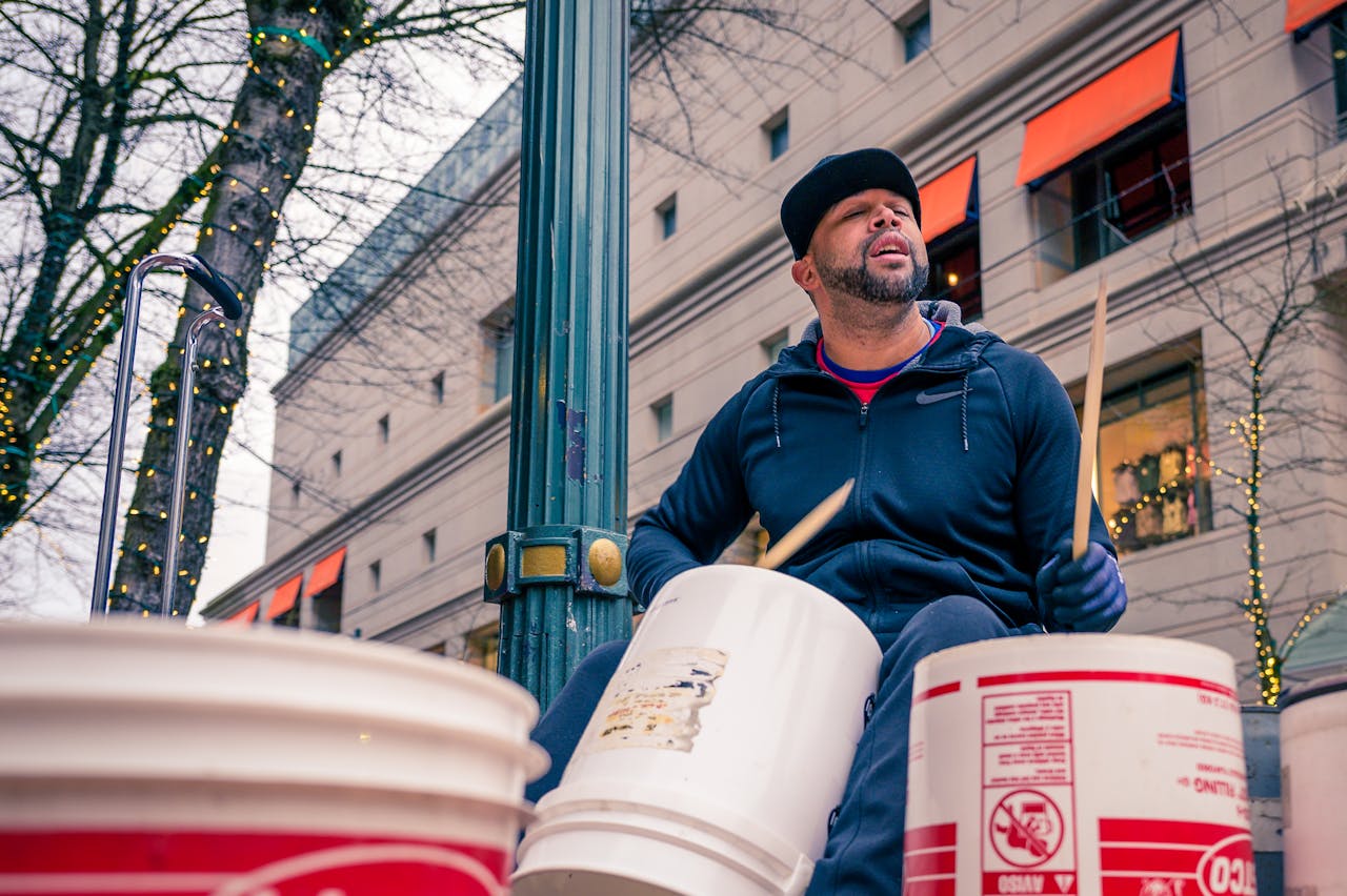 A street performer plays drums on buckets in a lively city environment during the daytime.