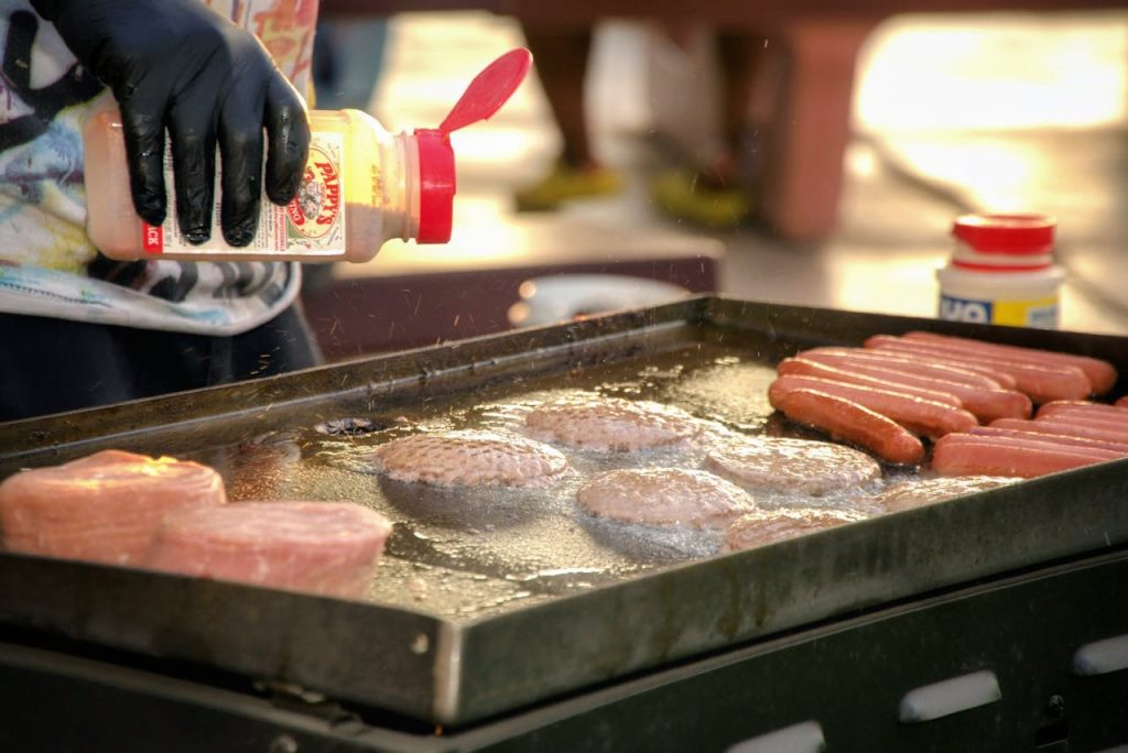 grilling-hot-dogs-and-burgers-at-outdoor-bbq-31901102 Chef seasoning hot dogs and burgers on a grill during an outdoor barbecue event.