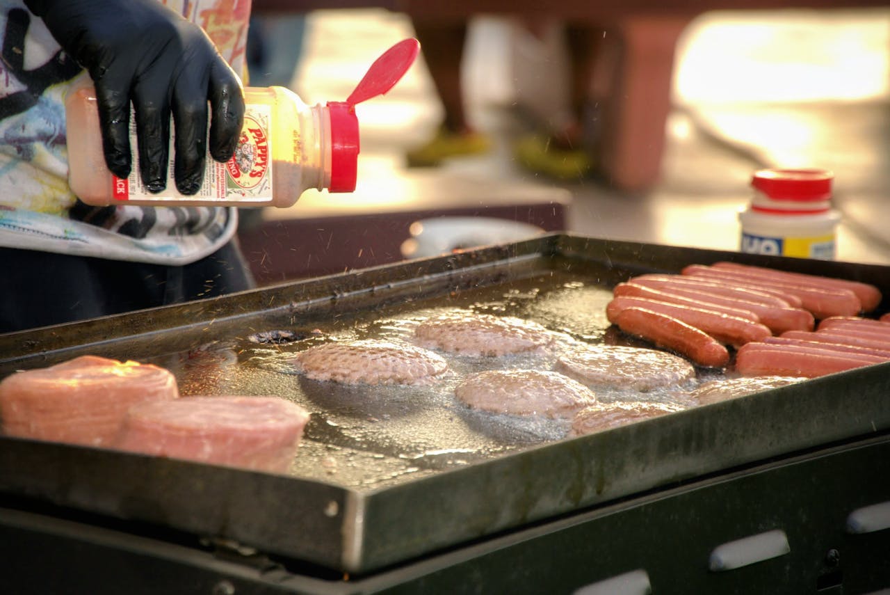 Chef seasoning hot dogs and burgers on a grill during an outdoor barbecue event.