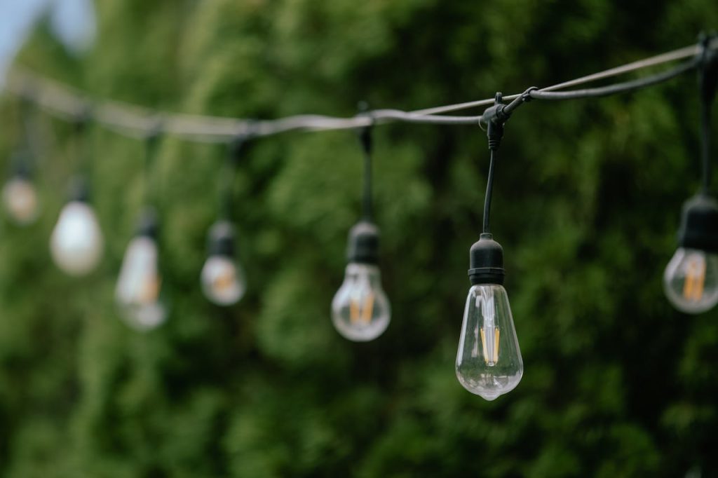 string-lights-in-garden-setting-31293623 Close-up of string lights hanging outdoors with green foliage in the background.